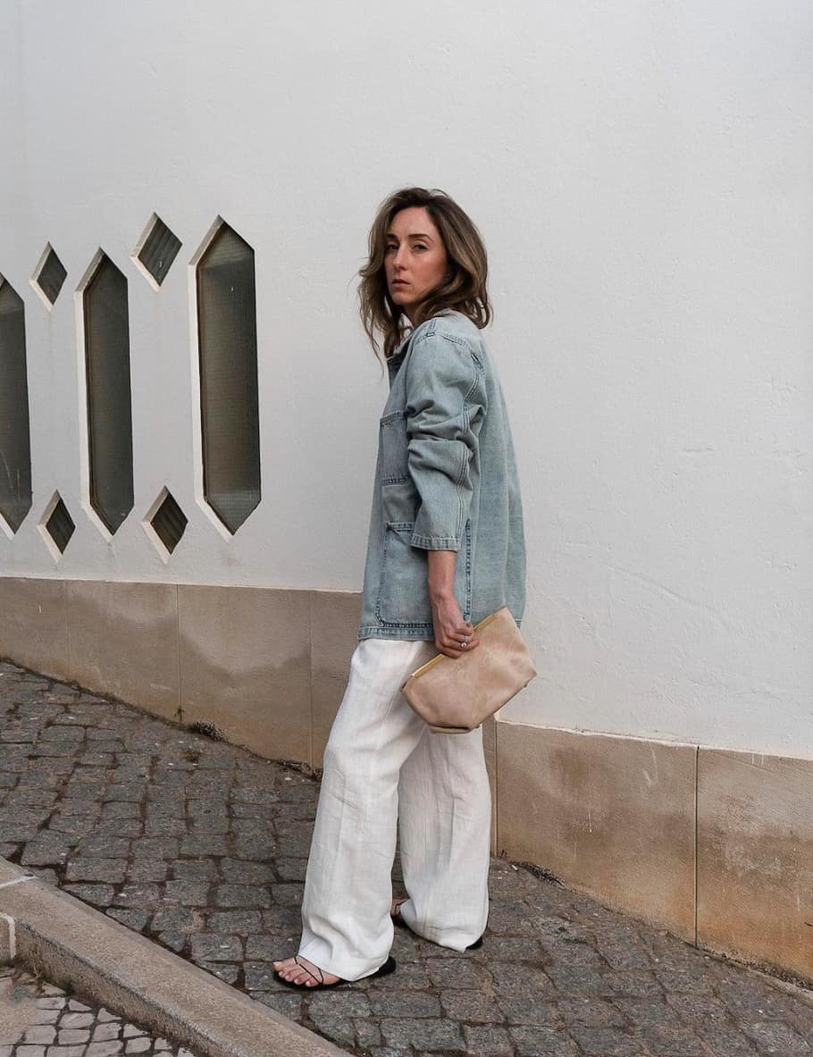 An image of a woman with white linen pants, a denim button-up, black sandals, and a tan clutch