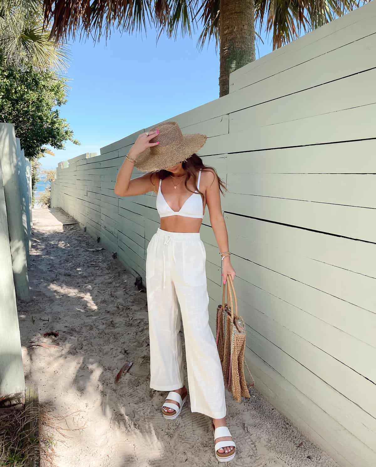 An image of a woman with white linen pants, chunky sandals, a white bikini top, and a straw hat and tote bag
