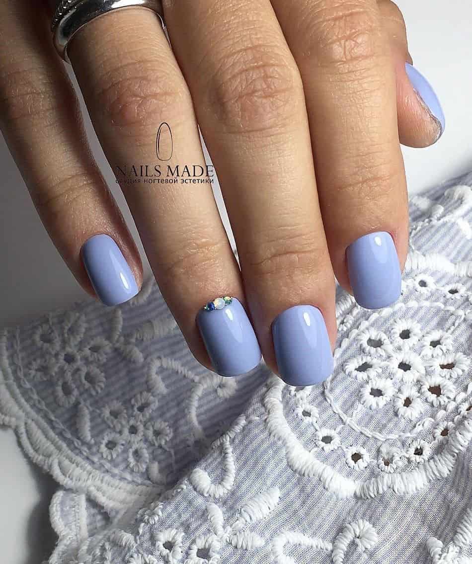 A hand with short square nails with periwinkle polish and gem accents