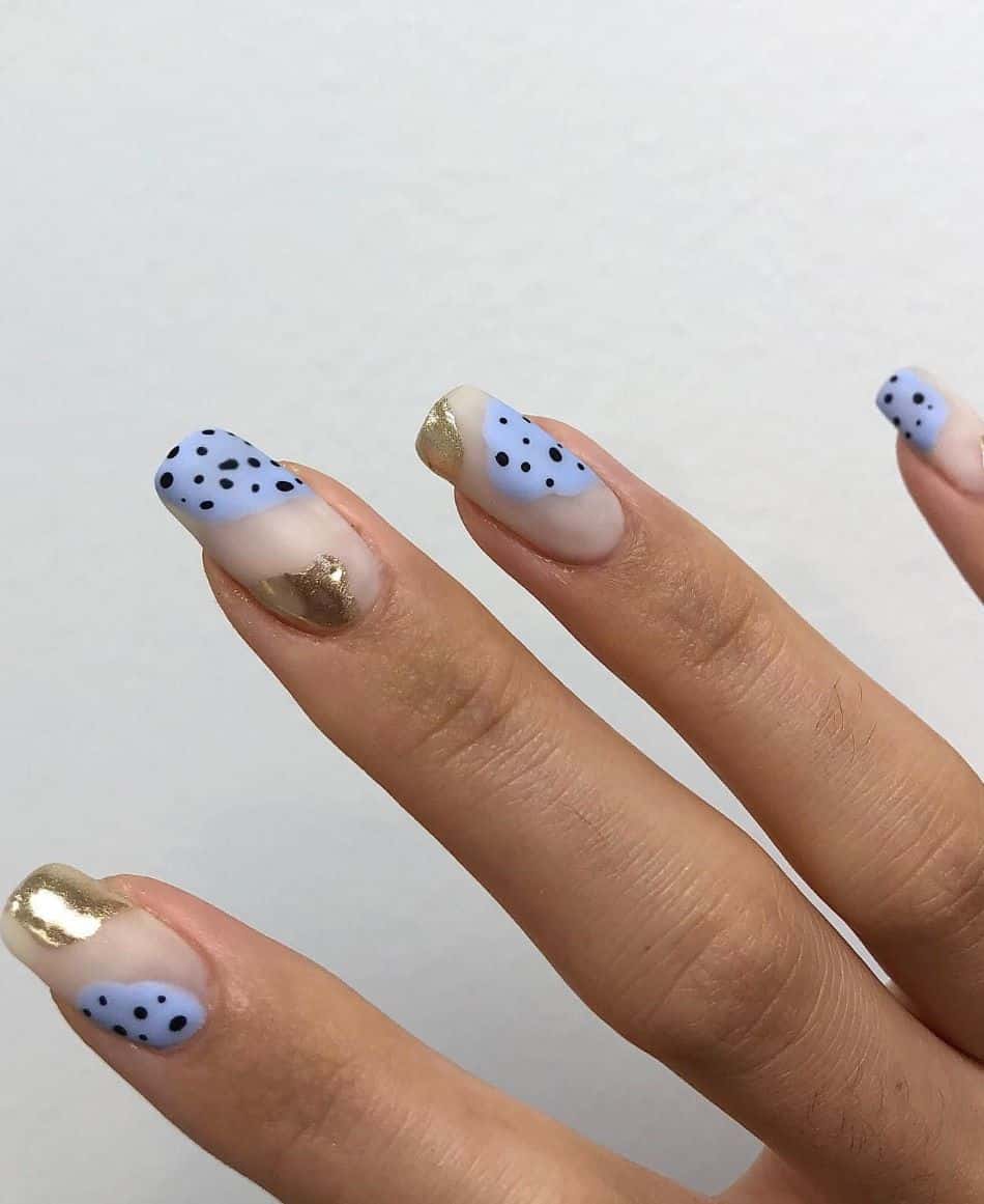 A hand with nude nails featuring periwinkle and gold polish with black dots