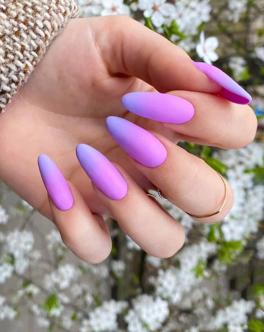 An image of a hand with long almond nails featuring a matte purple and indigo ombre