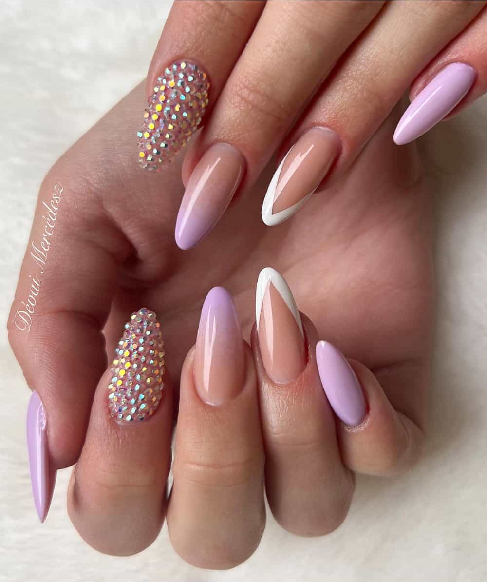 An image of a hand with multi patterned nails featuring white French tips, purple ombre, and gem accent nails