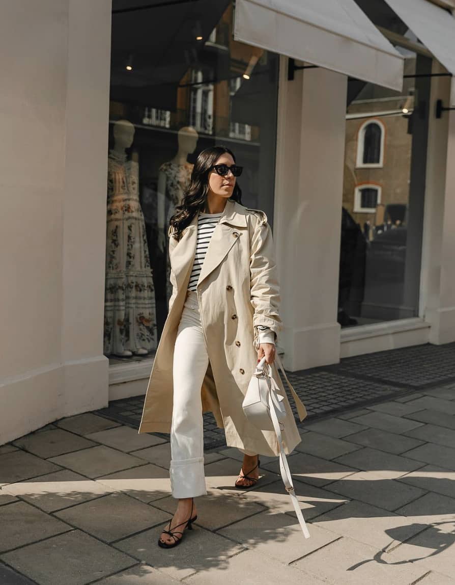woman wearing a tan trench coat over a striped top with off-white jeans and black heels