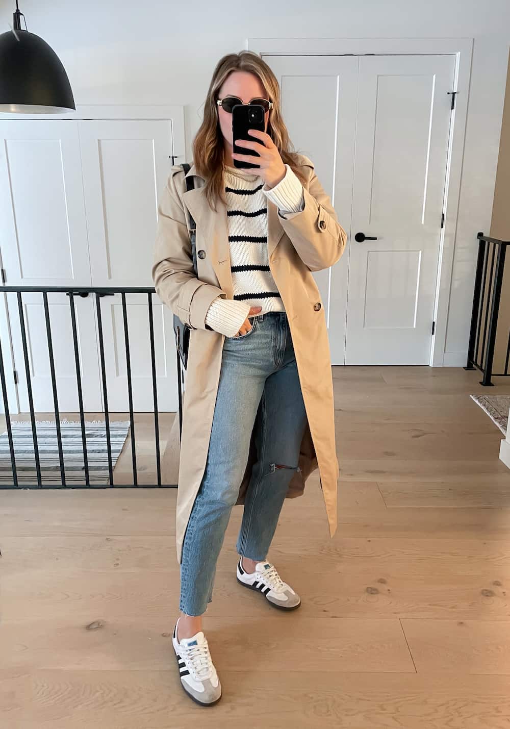 woman wearing a spring capsule wardrobe outfit with a tan trench coat, striped knit sweater, vintage wash jeans, and white sneakers