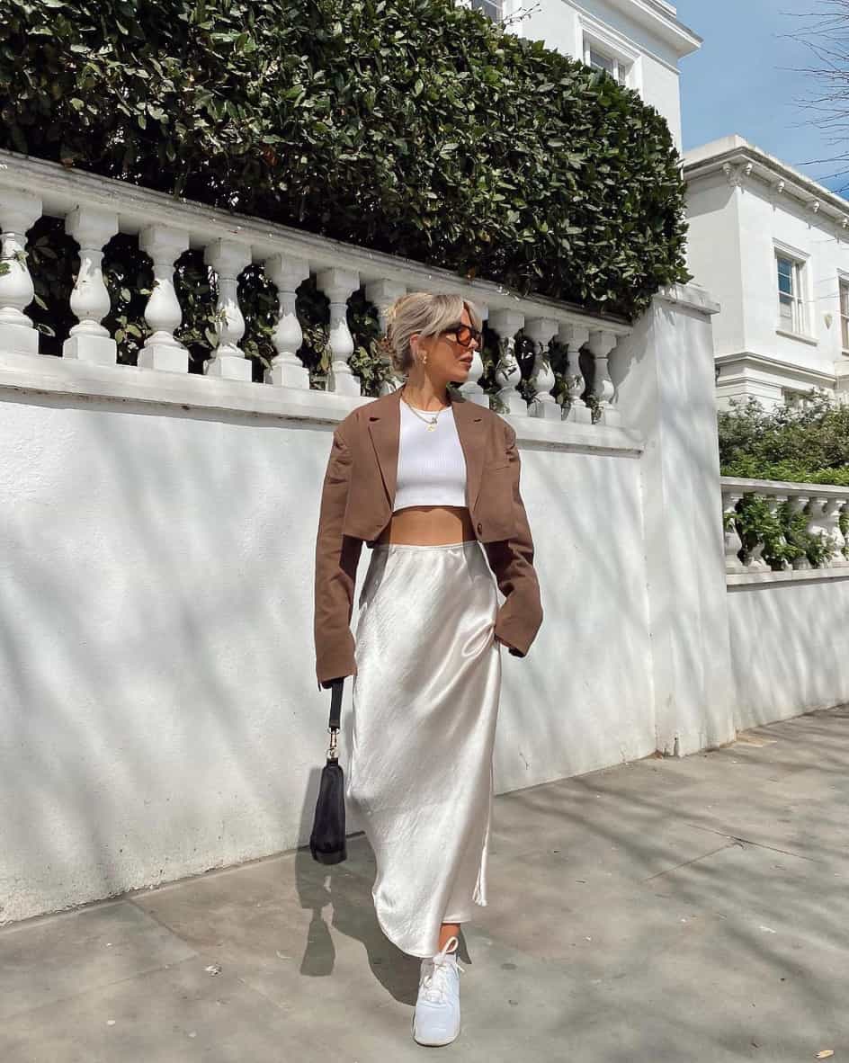 A woman wearing a white silk midi skirt with a cropped white tee, a cropped brown jacket, and white sneakers