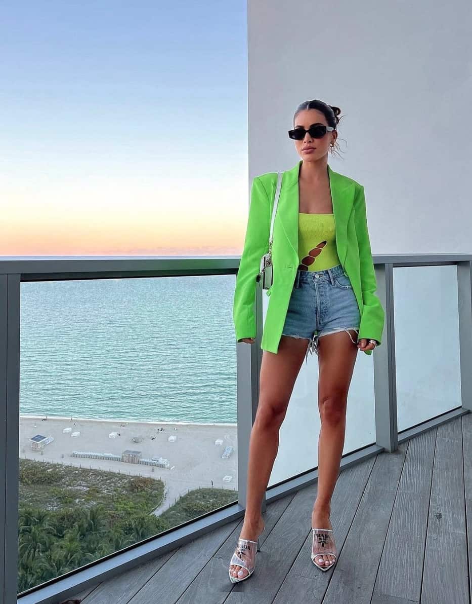 A woman wearing light blue denim shorts, a bright green bodysuit with cut-outs, a lime green blazer, and heels
