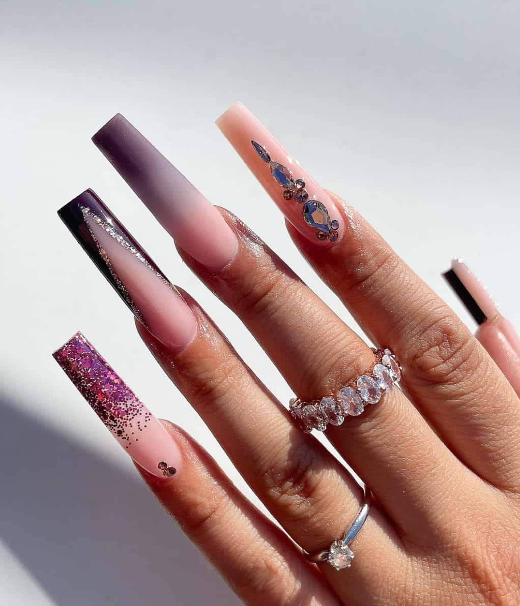 A hand with long nude and purple square nails with ombre, glitter, French tips, and gem accents