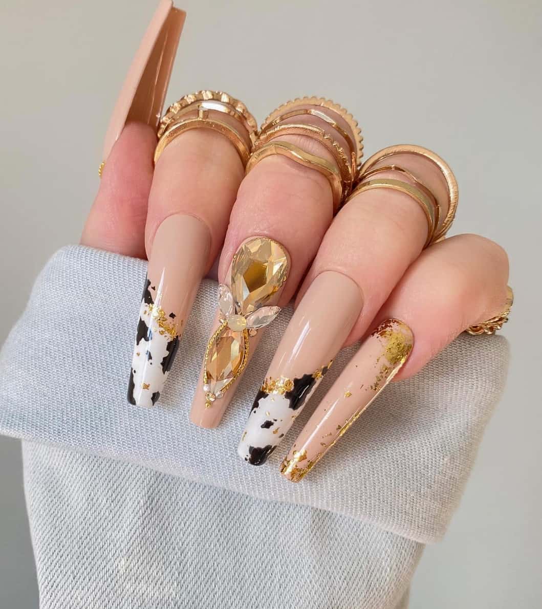 A hand with long coffin nails featuring a nude base polish with cow print accents, gold flake details, and gems