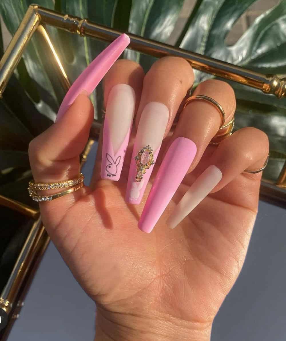 A hand with long coffin nails featuring nude and pink polish, French tips, Playboy Bunny logos, and gem accents