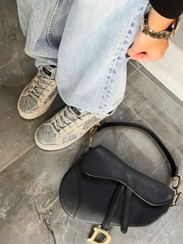 Womans legs in jeans wearing golden goose sneakers with a Dior saddle bag on the floor beside her feet