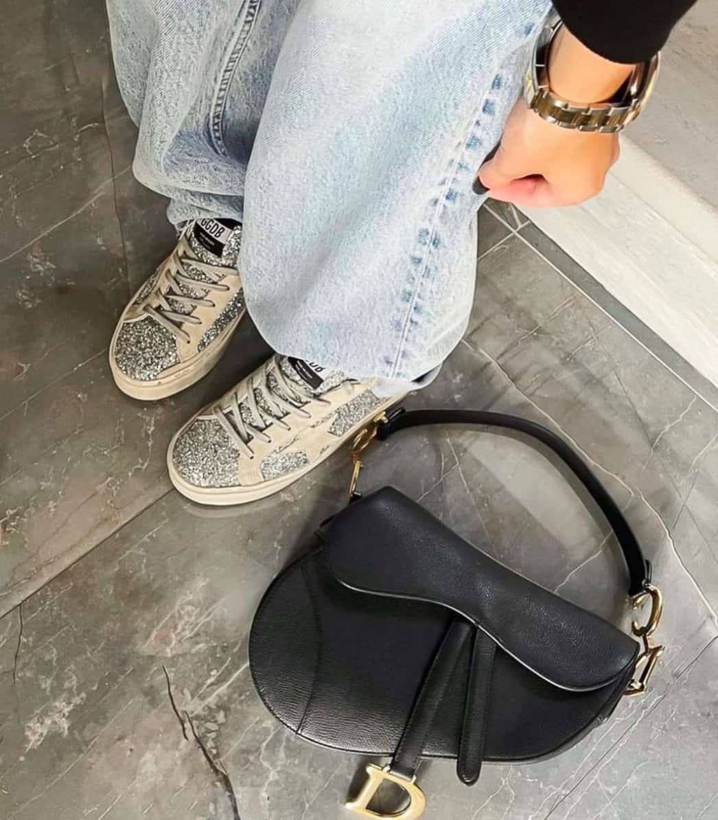Womans legs in jeans wearing golden goose sneakers with a Dior saddle bag on the floor beside her feet