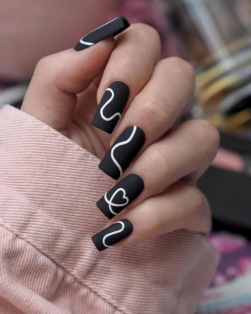 A hand with square matte black nails with white lines and a line heart