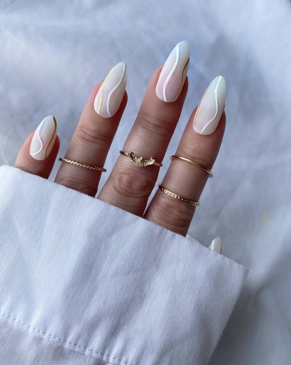 A hand with white matte almond nails featuring white and gold wavy accent lines