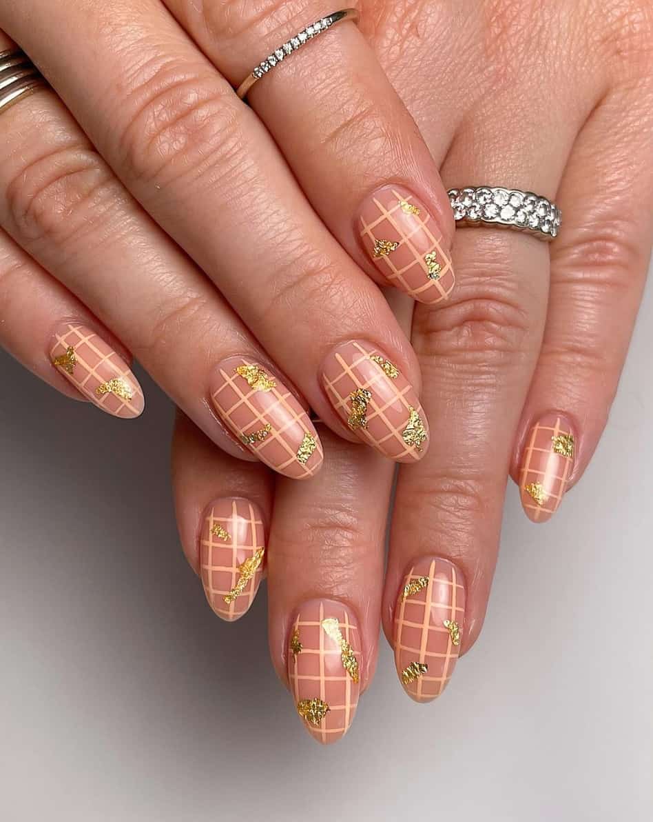 A hand with beige almond nails, pastel orange grids, and gold flakes