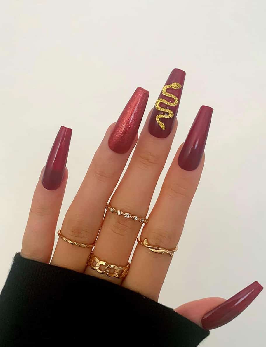 A hand with long coffin nails with dark red polish featuring a glittery red accent nail and a gold snake accent