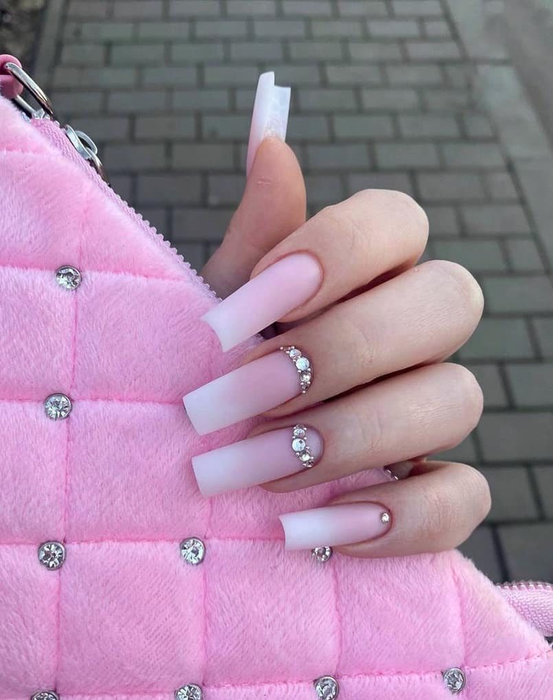 A hand with long square nails featuring white and pink ombre with a matte finish and gem accents