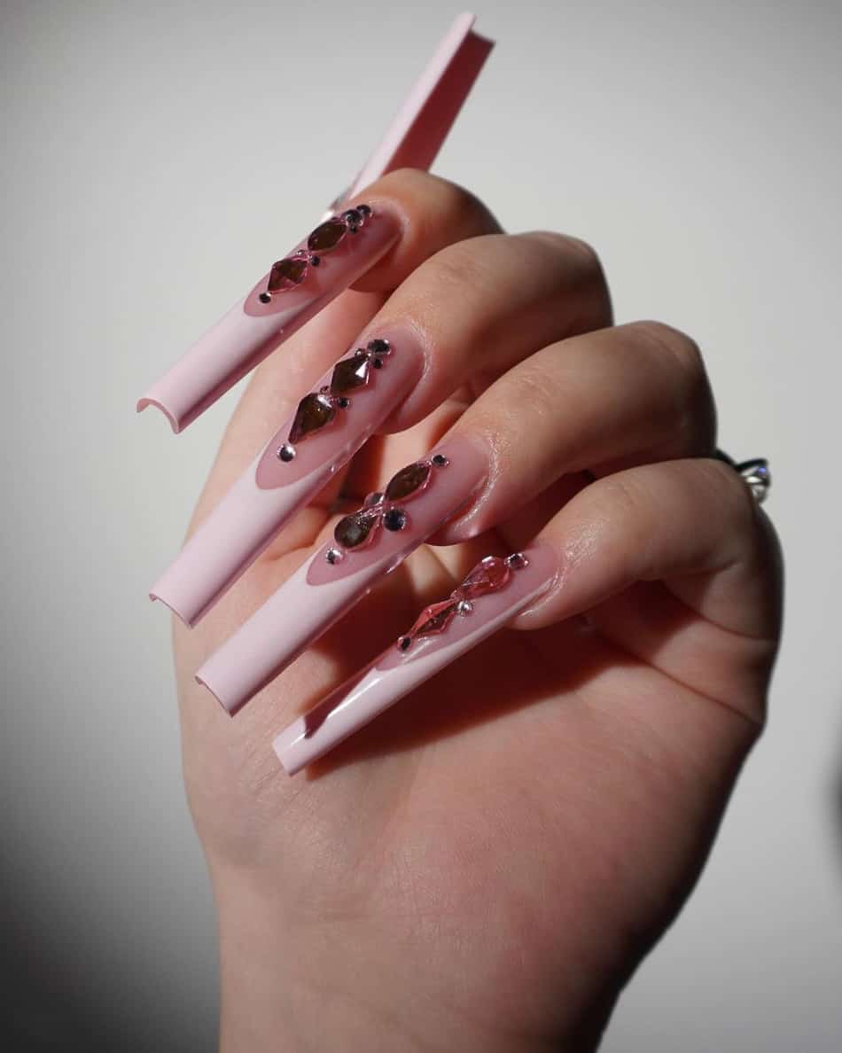 A hand with long coffin acrylic nails with a pink polish base, light pink French tips, and large gem accents