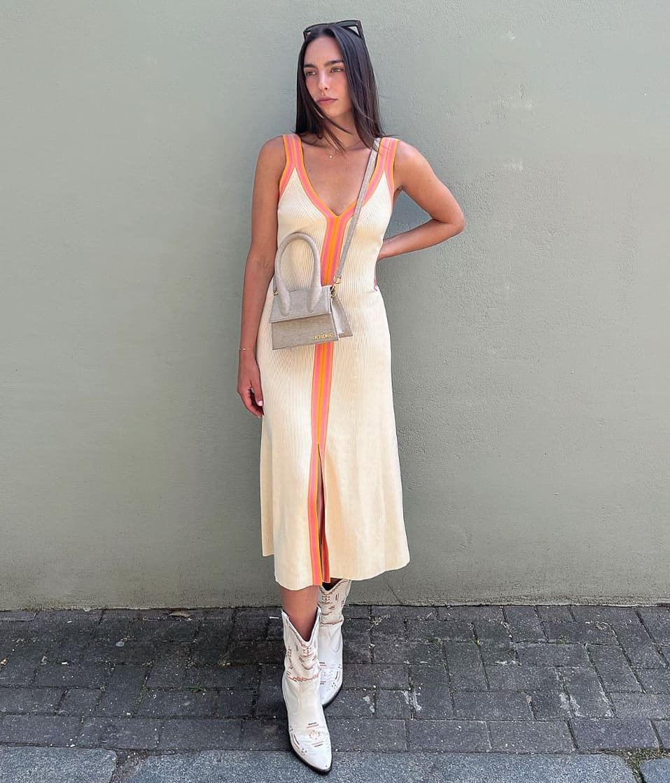 A woman wearing a cream midi summer dress with orange and pink trim, short silver cowboy boots, and a silver crossbody purse