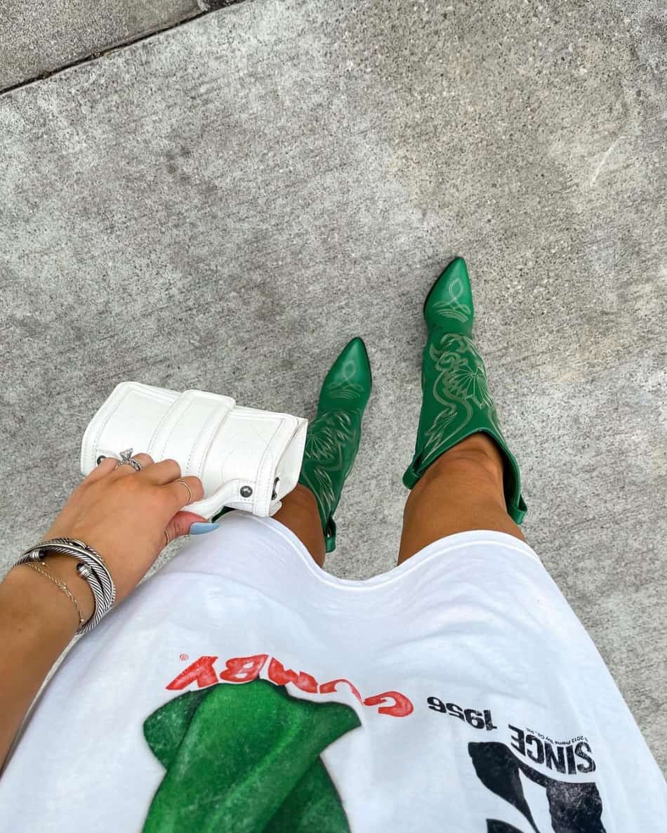 An overhead view of a woman wearing an oversized graphic tee and green cowboy boots holding a mini white handbag