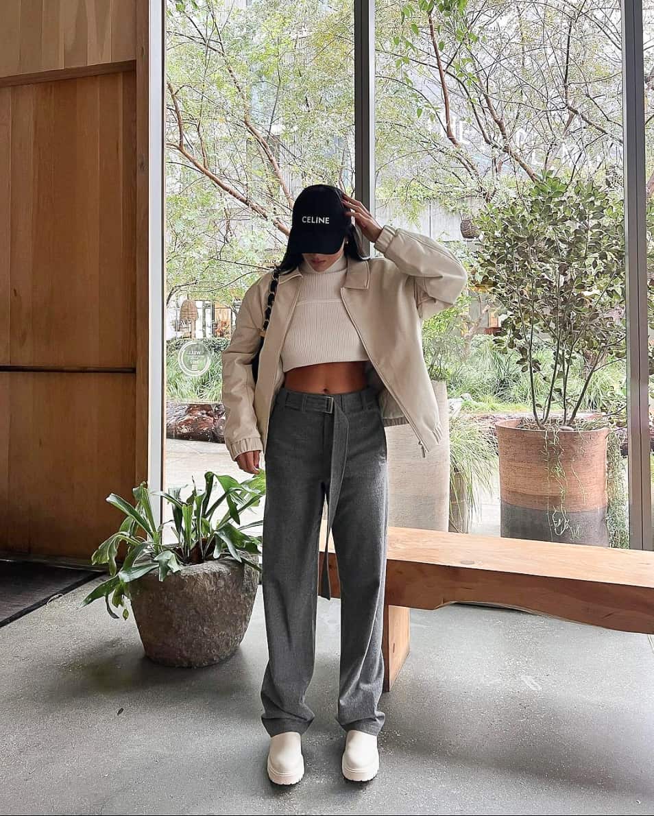 A woman wearing dark grey pants with a white crop top, a beige jacket, and beige boots