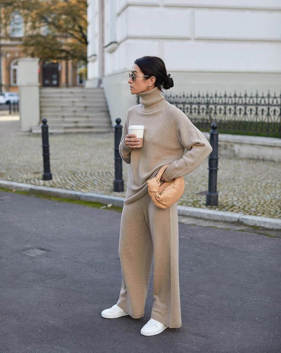 A woman wearing a matching beige set featuring a turtleneck sweater and wide leg trousers and white sneakers