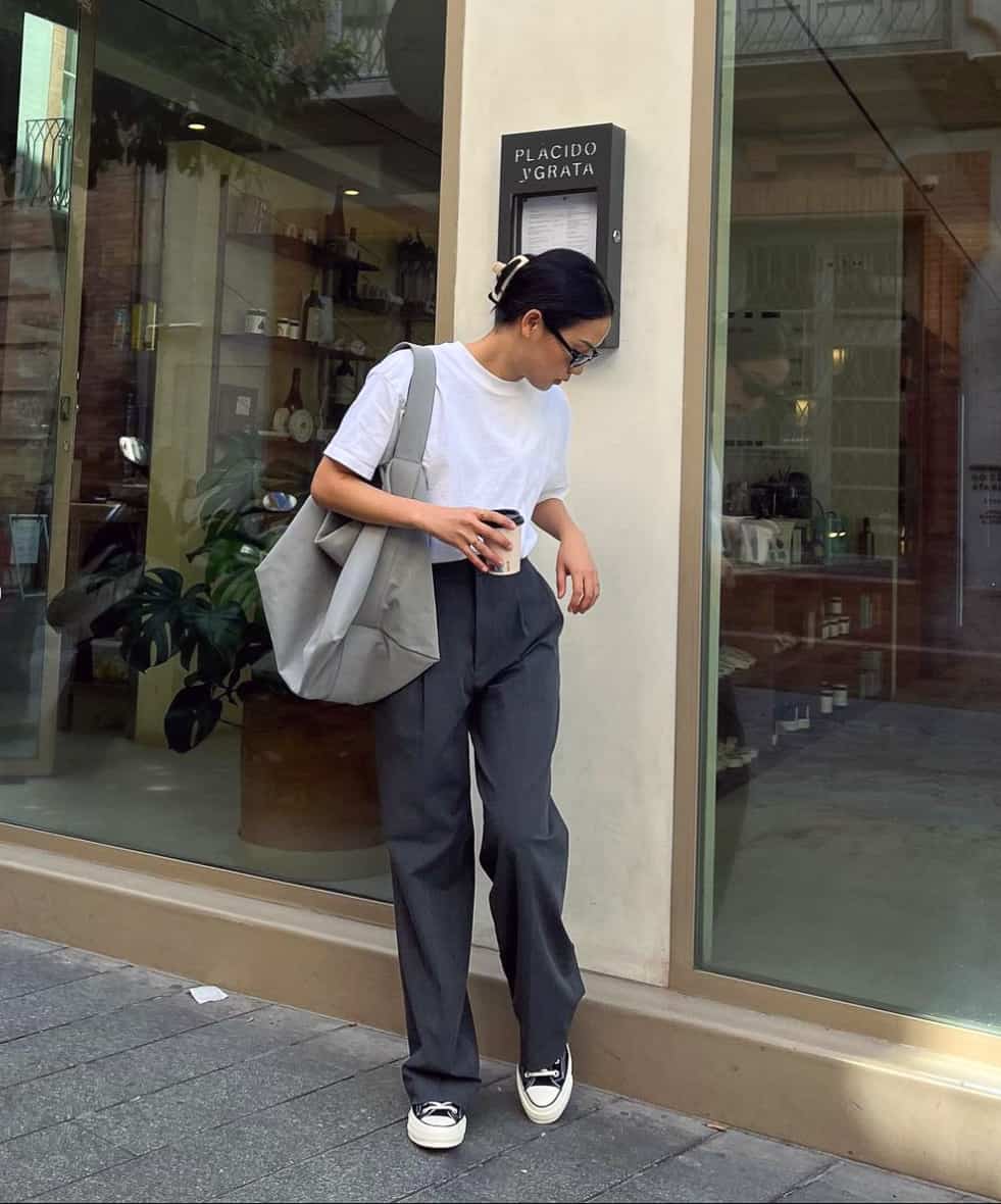 A woman wearing dark grey trousers, a tucked-in plain white tee, Converse sneakers, and a large grey tote bag