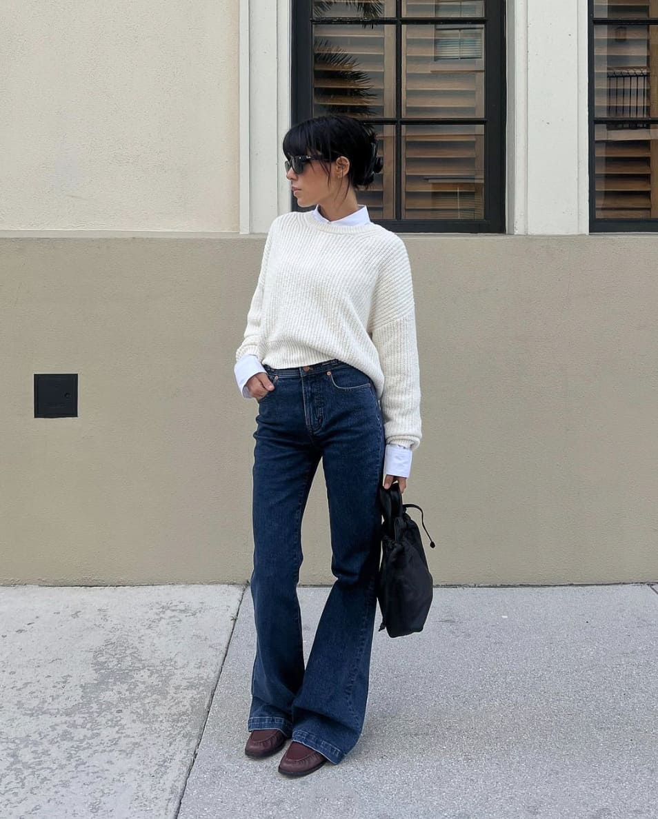 A woman wearing dark blue flared jeans with a cream-colored sweater over a white button-up, accessorized with a black handbag, brown boots, and black sunglasses