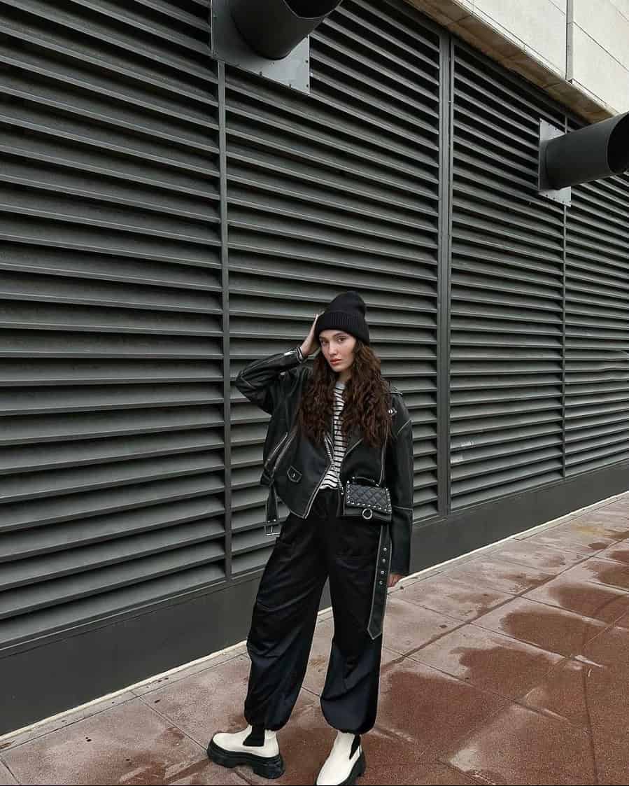 A woman wearing baggy black pants and a black and white striped tee with a black leather jacket, black and white platform Chelsea boots, and a black beanie