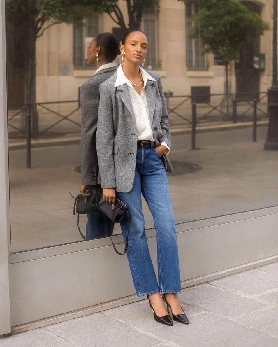 A woman wearing a white button-up tucked into belted blue jeans with a grey blazer, black pumps, and a black handbag