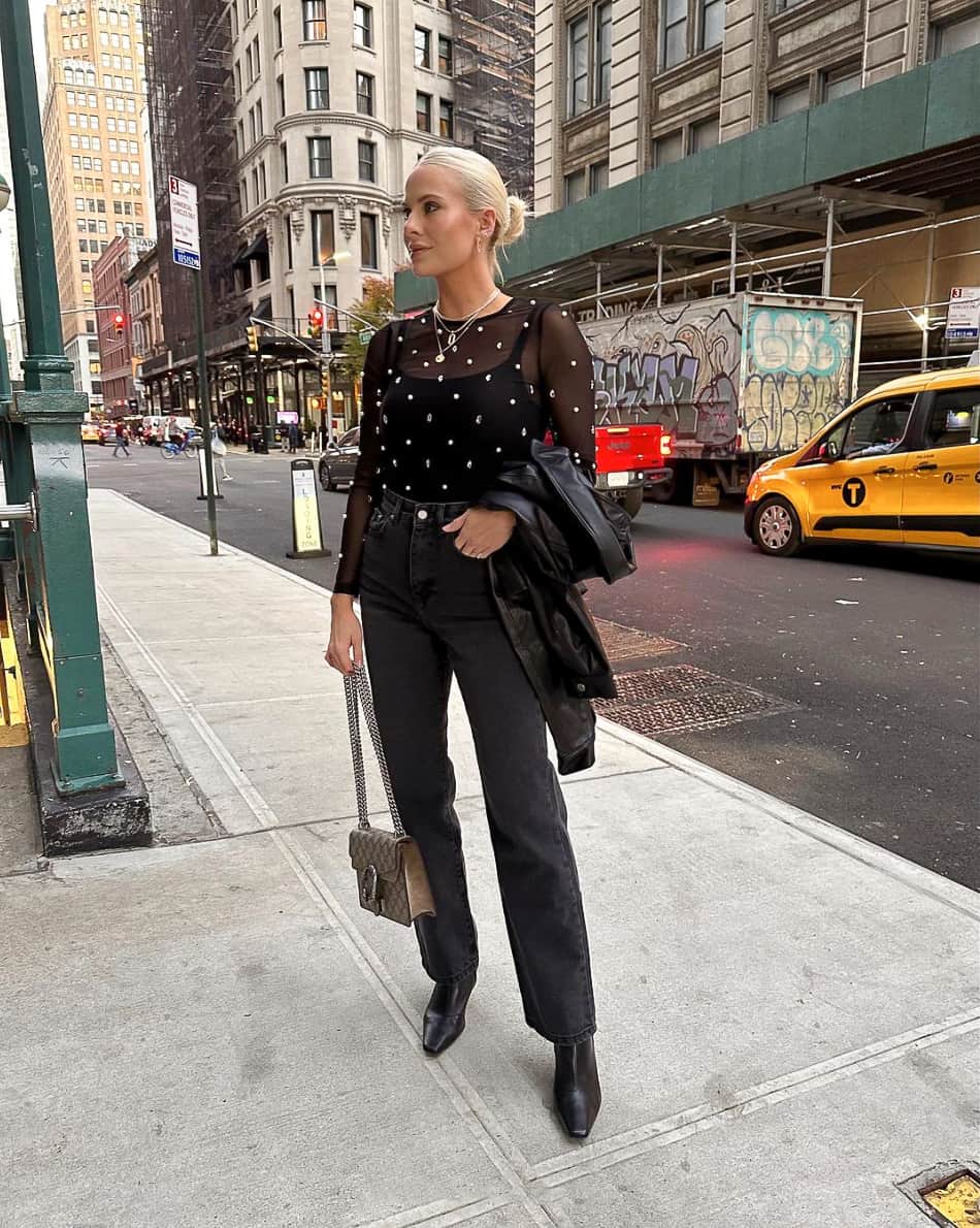 A woman wearing black jeans with a black tank top, black heeled boots, and a sheer long-sleeved top with gem accents