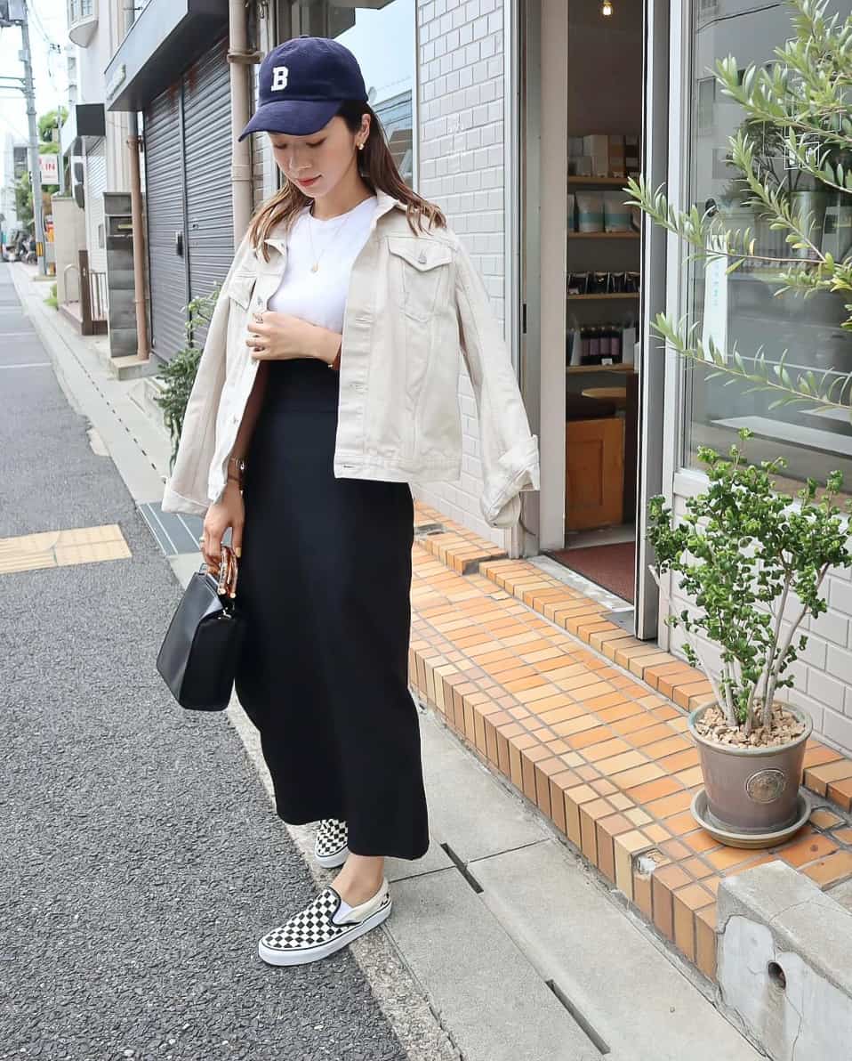 A woman wearing a black maxi skirt, a white cropped tee, and an oatmeal-colored denim jacket with black and white checkered slip-on sneakers