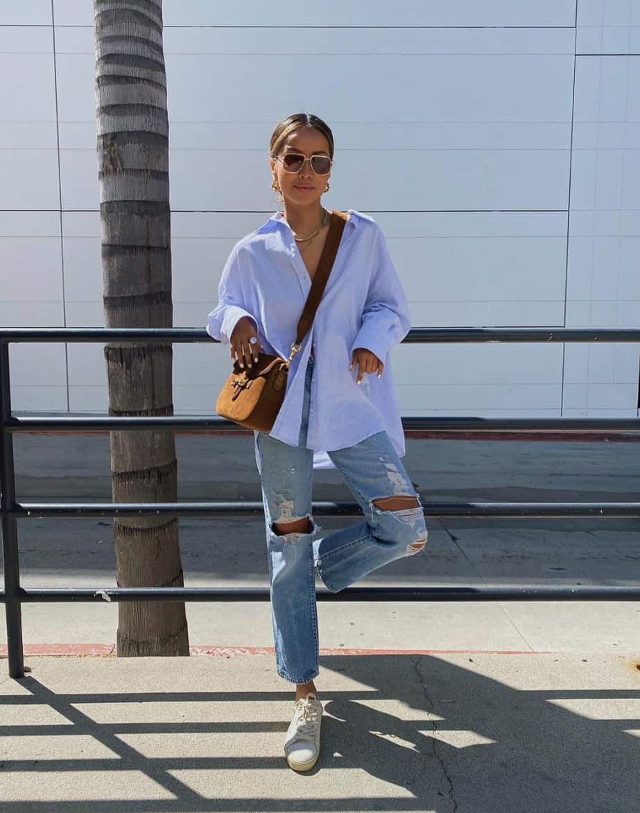 A woman dressed for a theme park with ripped blue jeans, a light blue button-up, white sneakers, and a cognac crossbody bag