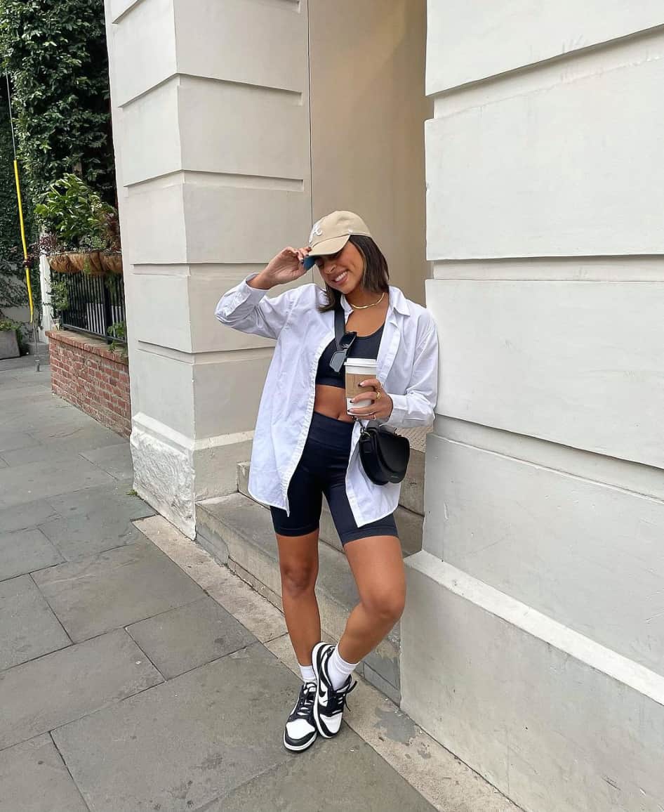 A woman dressed for a theme park wearing a black crop top, black running shorts, a white button-up, black and white sneakers, and a beige baseball cap