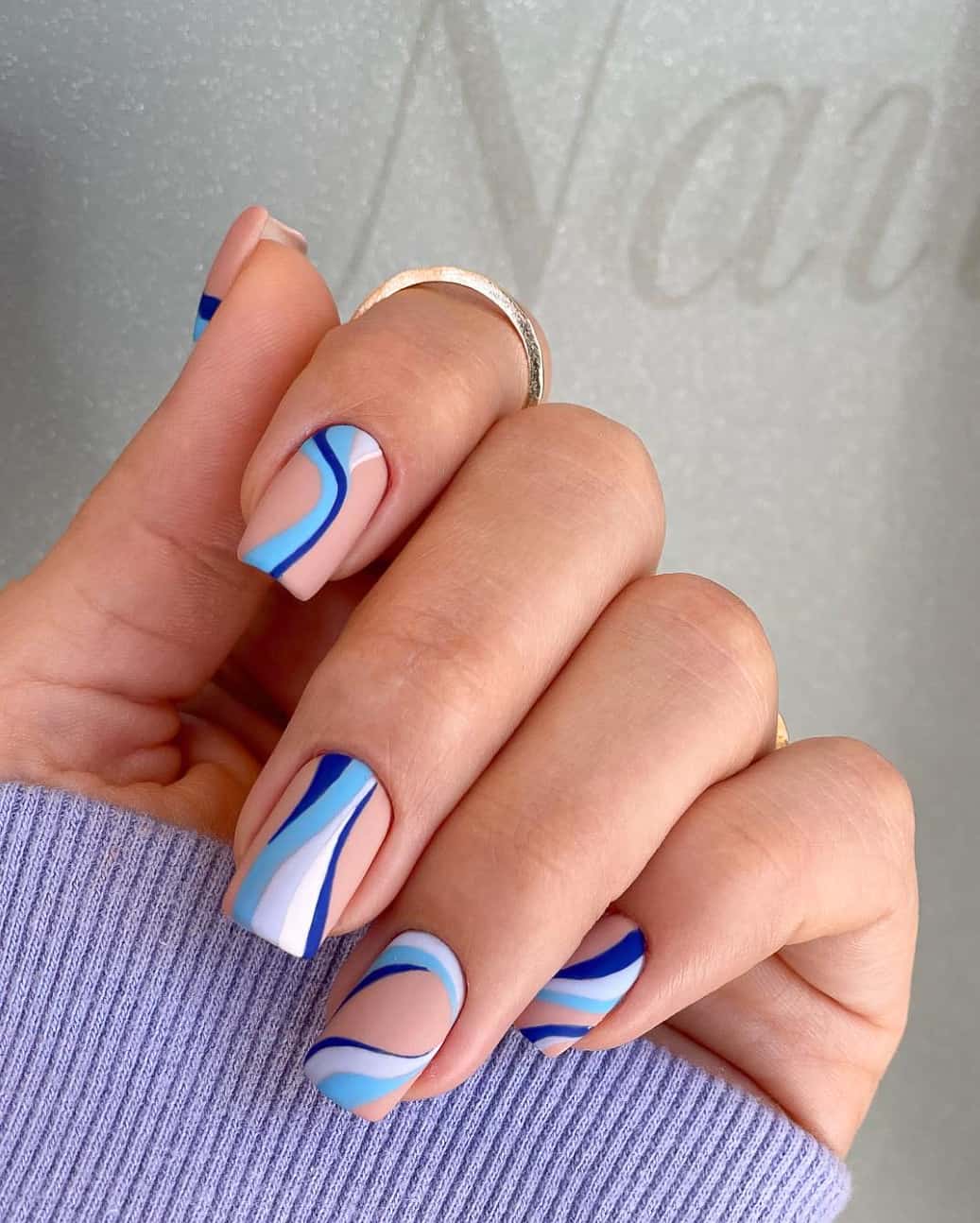 A hand with matte nude square nails featuring swirls in multiple shades of blue and white