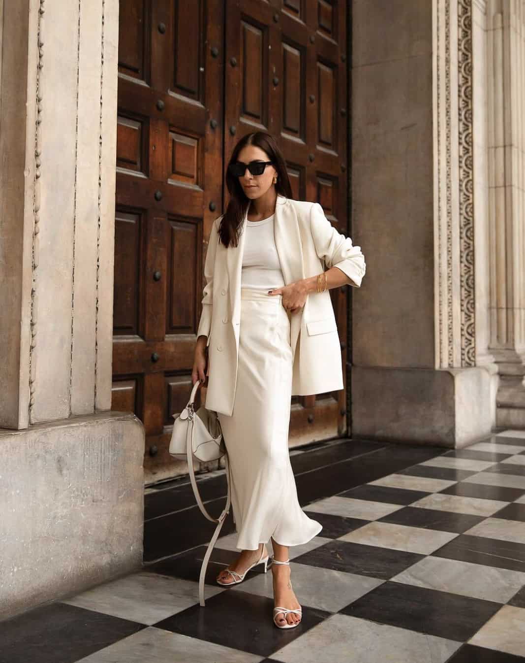 A woman wearing a full white outfit consisting of a silk midi skirt, tank top, blazer, strappy heels, and a handbag