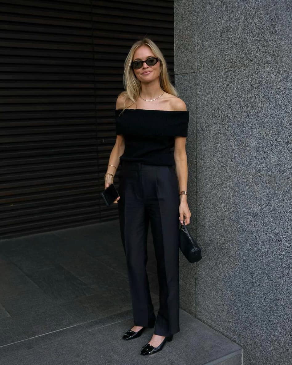 A woman wearing black pleated pants with an off the shoulder black top, low black heels, and a black clutch