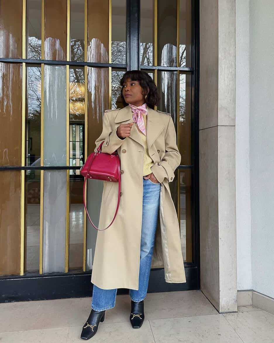 A woman wearing blue jeans with a yellow top, a classic long trench coat, black heeled boots, and a red handbag