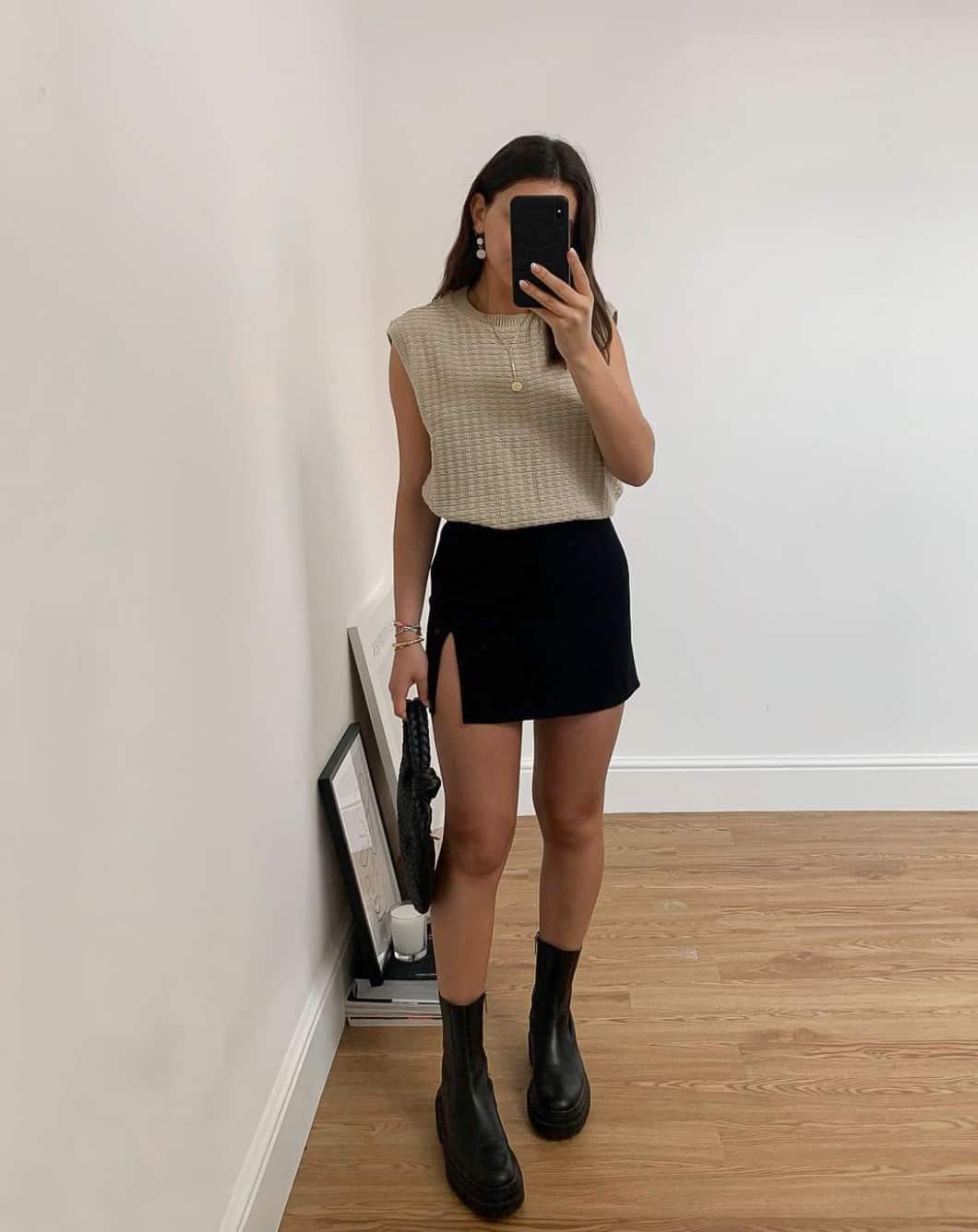 A woman wearing a slitted black mini skirt with a light beige sweater vest and black ankle boots