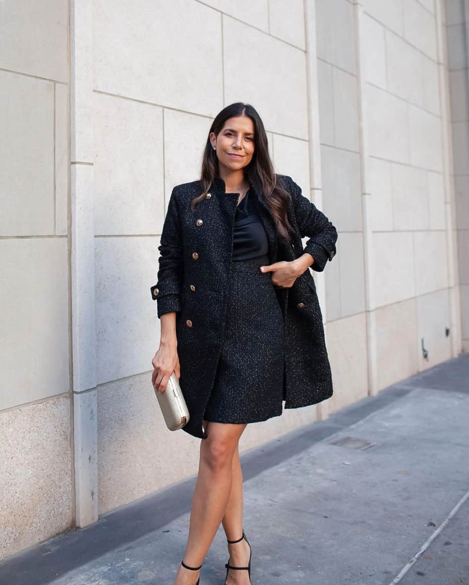 A woman wearing a black tweed skirt and jacket set with a silk blouse, strappy black heels, and a metallic clutch