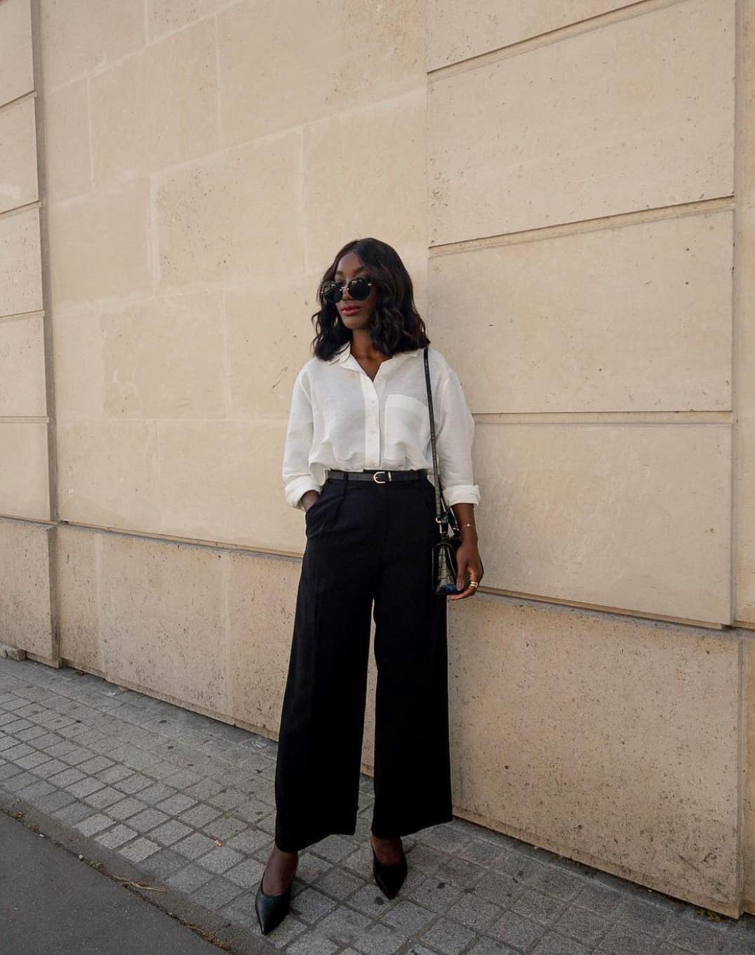 A woman wearing wide-leg black trousers with a white button-up tucked in, black heels, a black purse, and sunglasses