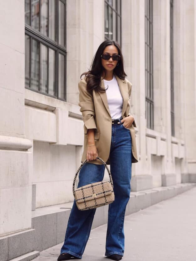 A woman wearing relaxed fit dark blue jeans with a white top, a beige blazer, and a beige and black plaid handbag