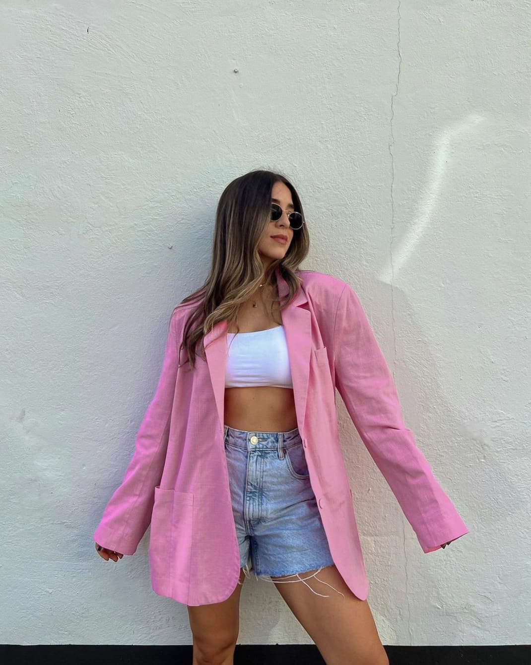 A woman wearing light blue denim shorts, a white bandeau top, and an oversized pink blazer with sunglasses