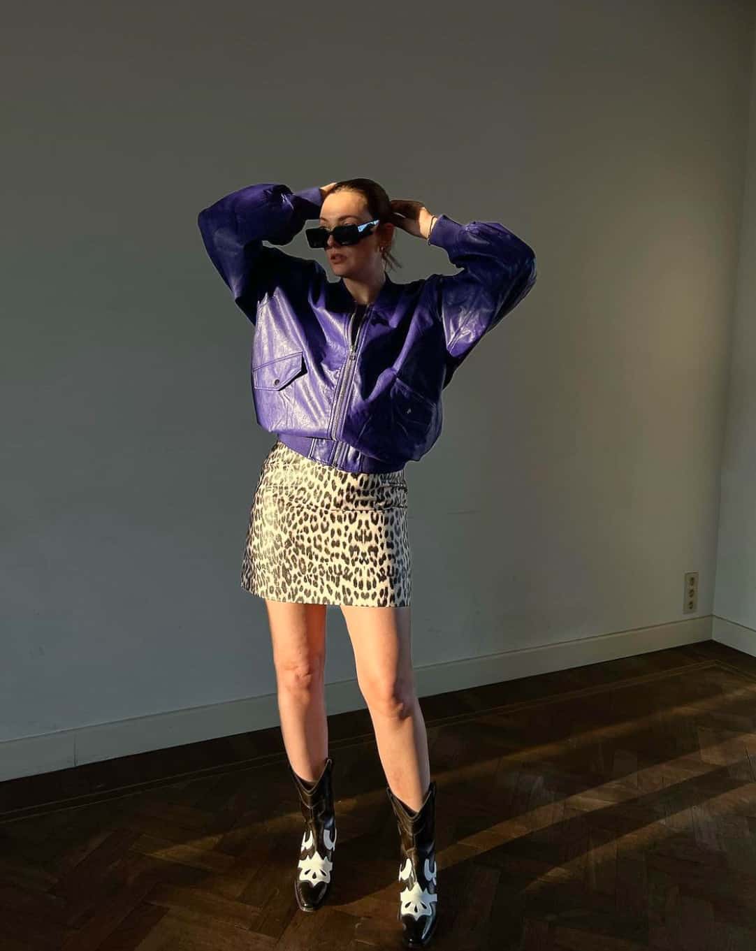 A woman wearing a leopard print mini skirt with black and white cowboy boots and a bright purple bomber jacket