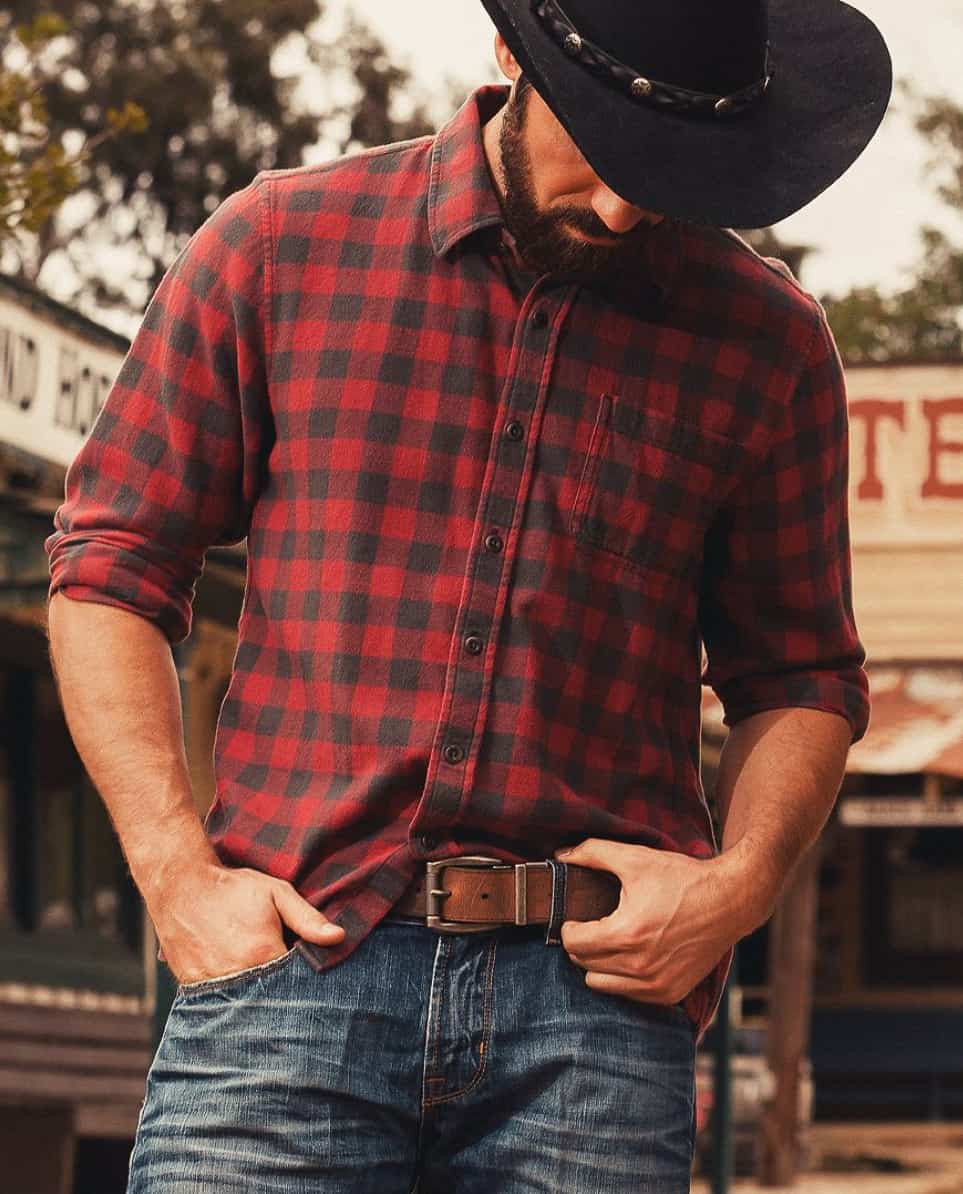 A man wearing blue jeans and a buttoned red flannel shirt with a brown belt and a black cowboy hat