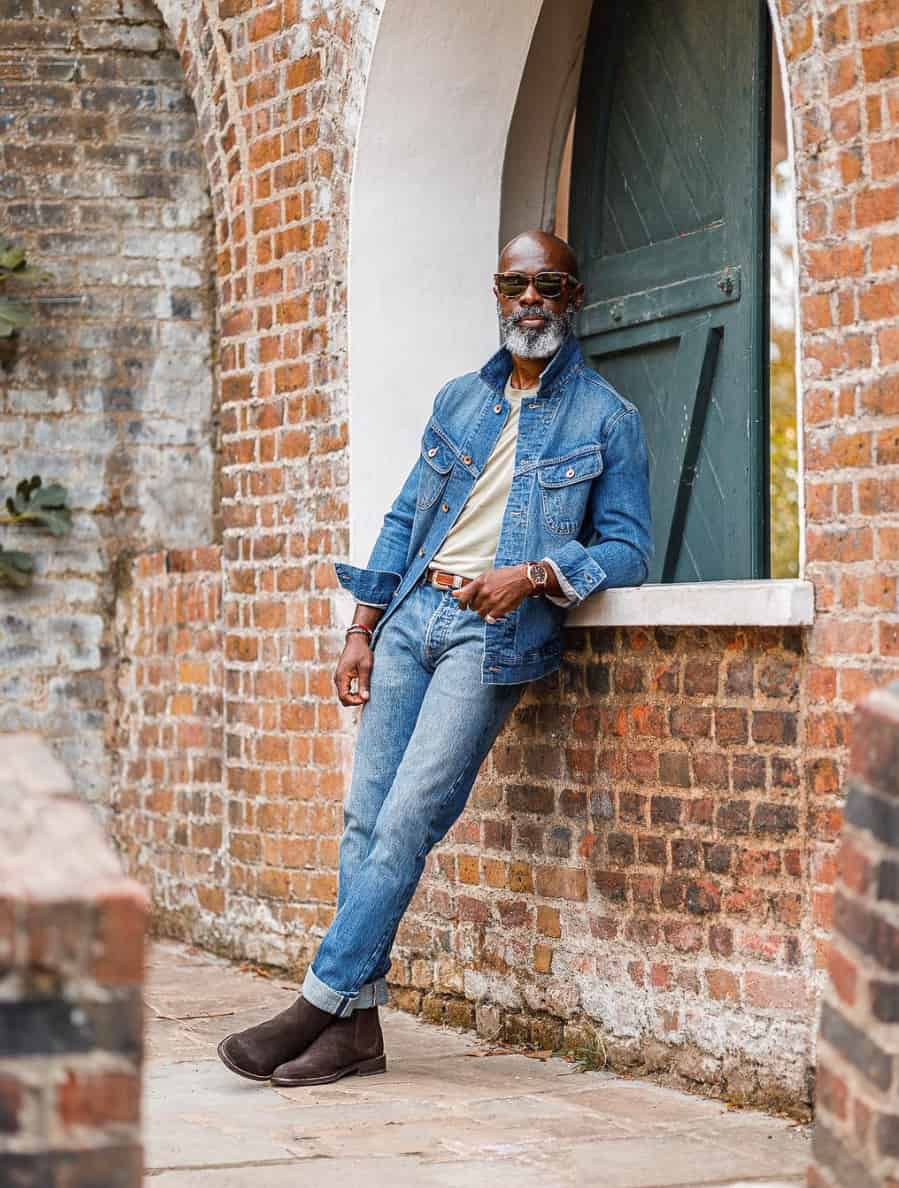 A man wearing blue jeans with a matching blue denim jacket, a plain tee, and dark brown suede boots