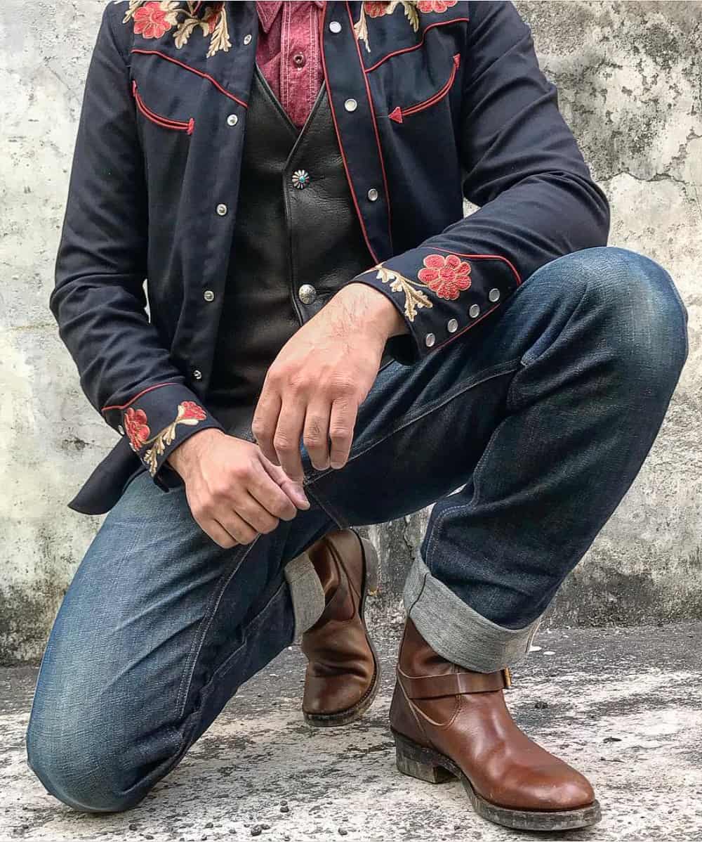A man wearing blue jeans with brown boots, a red button-up, a buttoned leather vest and an embroidered Western shirt on top