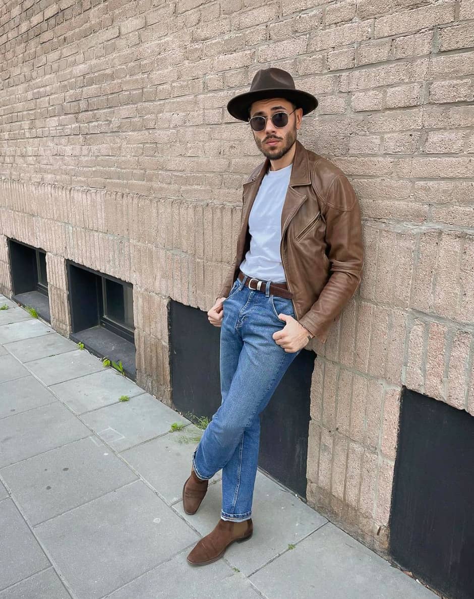 A man wearing blue jeans with brown boots, a white shirt, and a brown leather jacket with a dark brown fedora