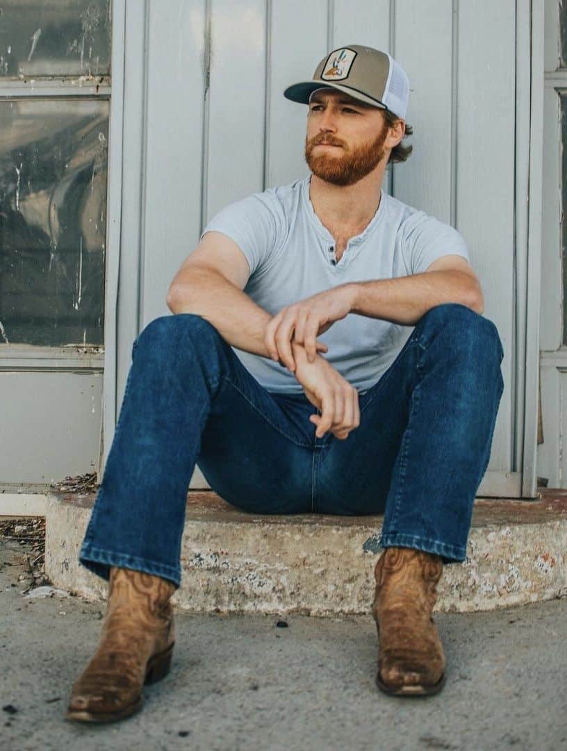 A man wearing blue jeans with a grey tee, brown cowboy boots, and a baseball cap