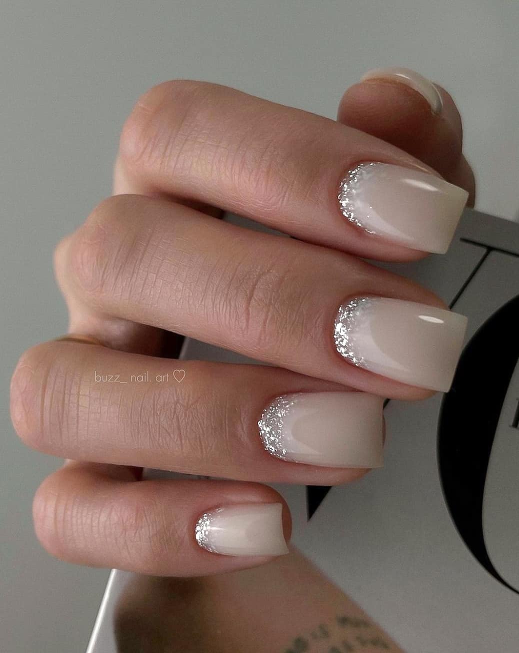 A hand with short square nails with a milky white base and silver glitter accents along the bottom