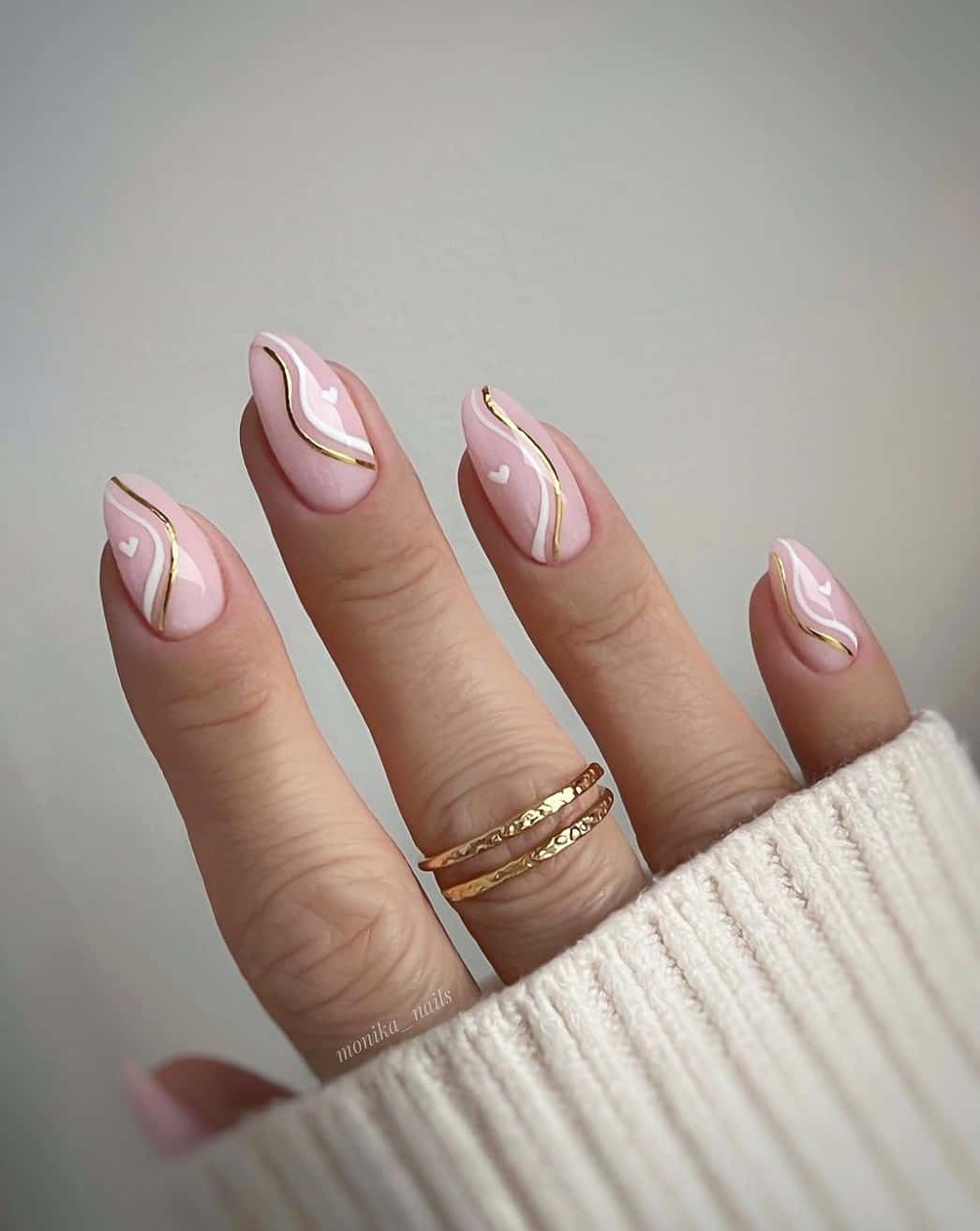 A hand with medium length nude almond nails with white wavy lines, gold wavy lines, and tiny white hearts